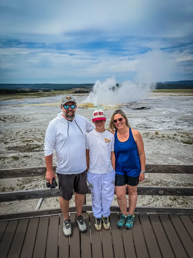 Amy Hartt smiling with her husband, Simon, and their son, Josiah, standing together outdoors in front of a steaming geyser basin and a wide blue sky.
